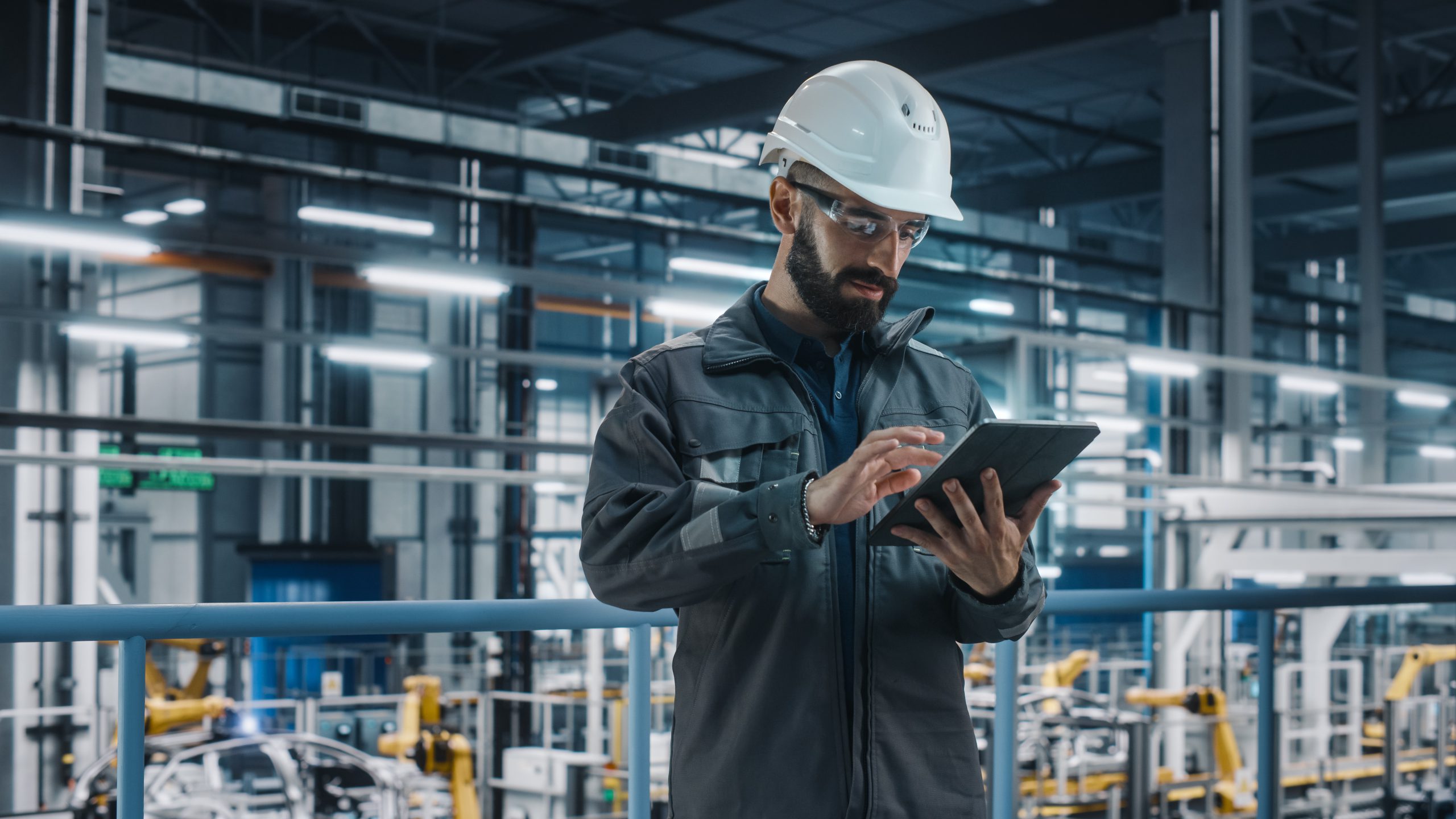 Portrait of Automotive Industry Engineer in Safety Glasses and Uniform Using Laptop at Car Factory Facility. Professional Assembly Plant Specialist Working on Manufacturing Modern Electric Vehicles.